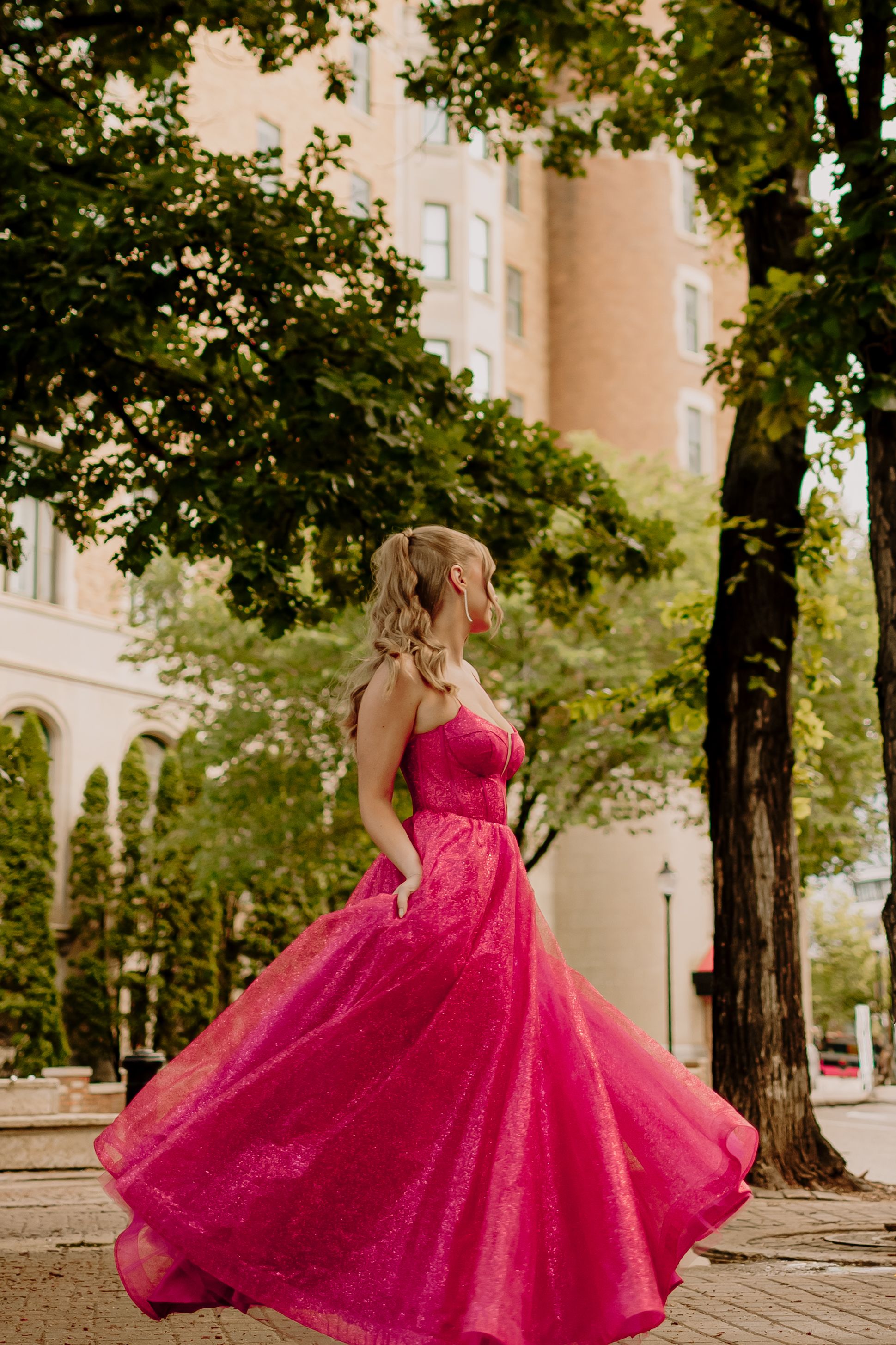 A vibrant hot pink ball gown twirls gracefully against a backdrop of trees and stone architecture.