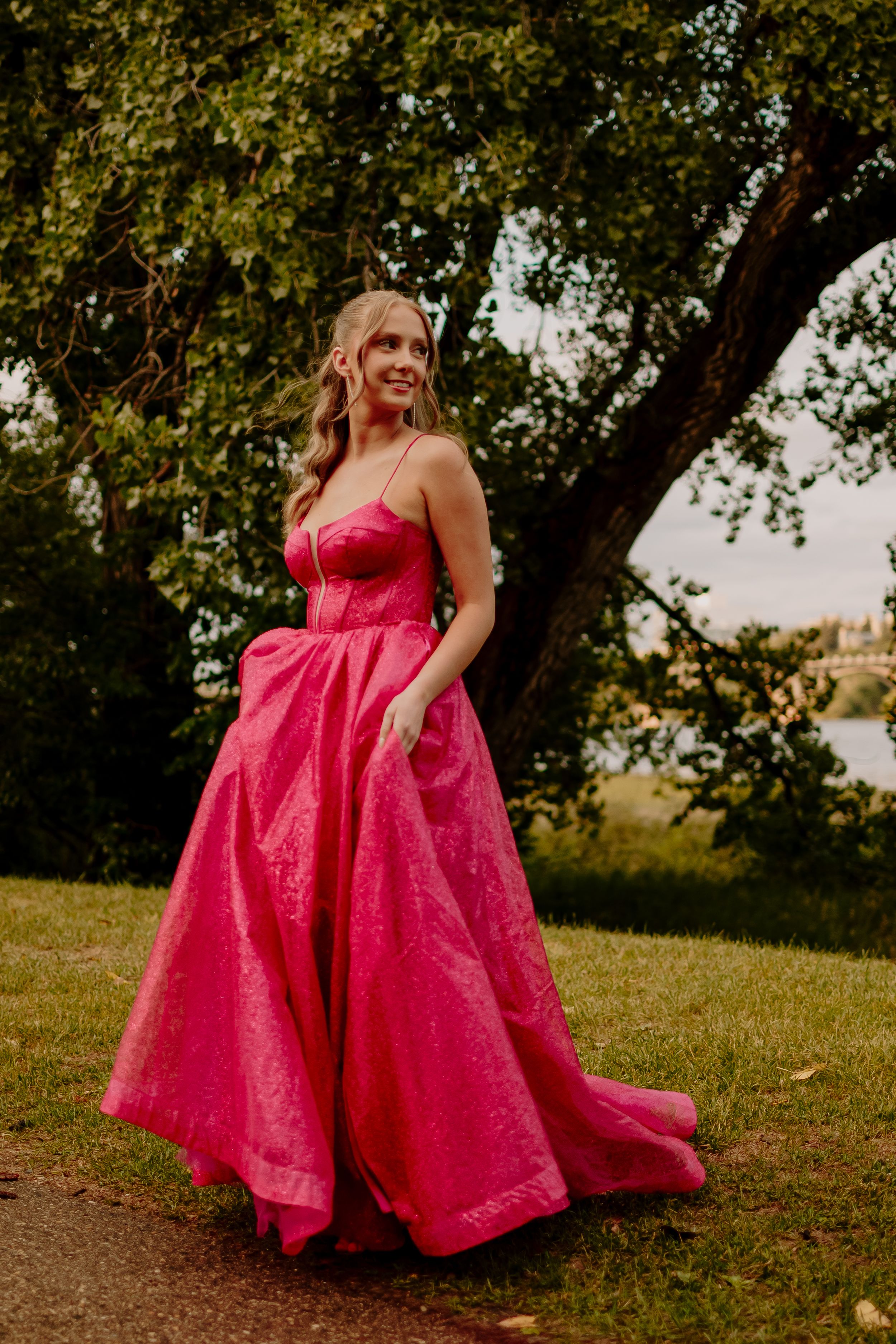 A flowing hot pink ball gown dress twirls gracefully under a tree in an outdoor setting.
