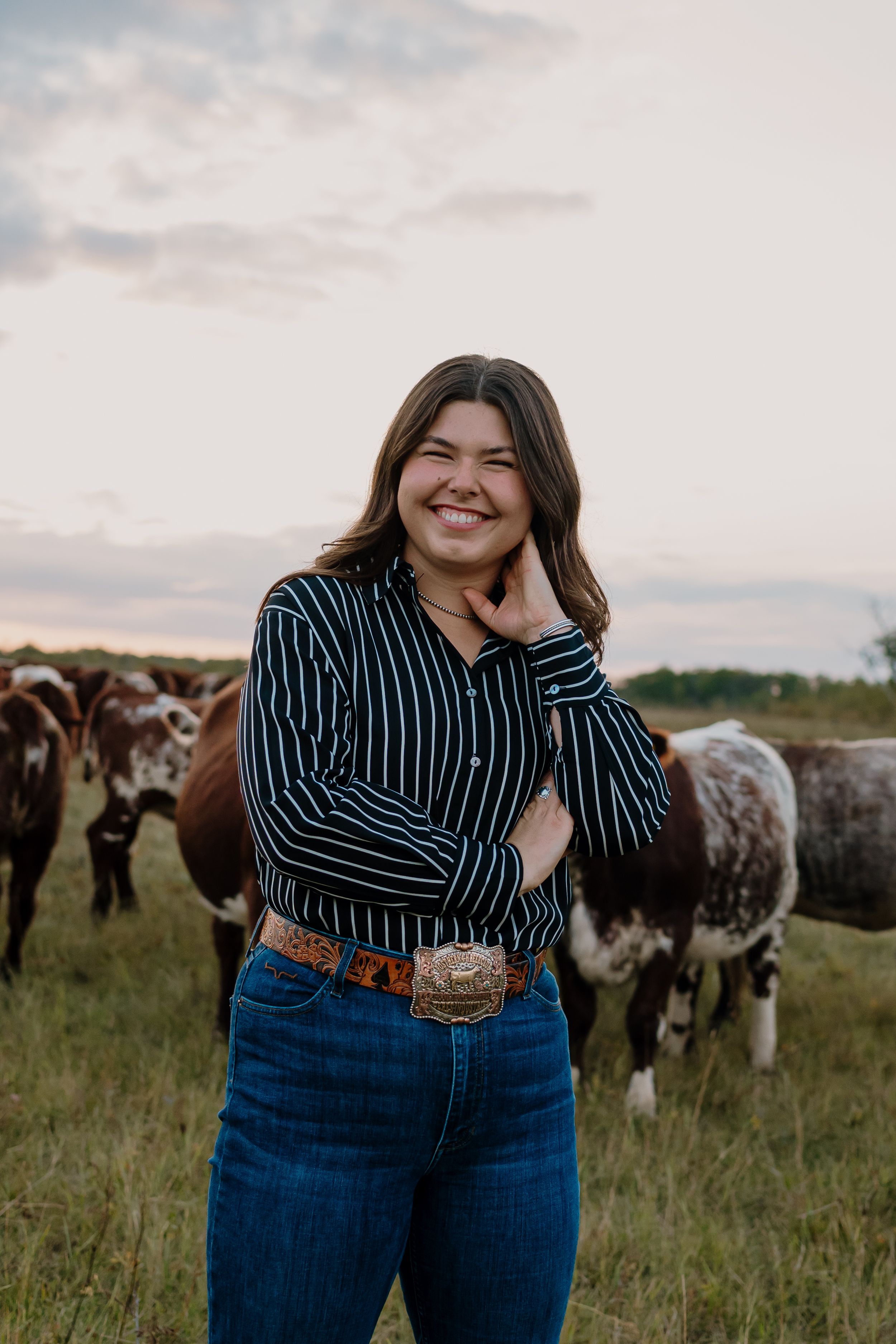 A person in a striped shirt and jeans poses in a pasture with longhorn cattle behind them.