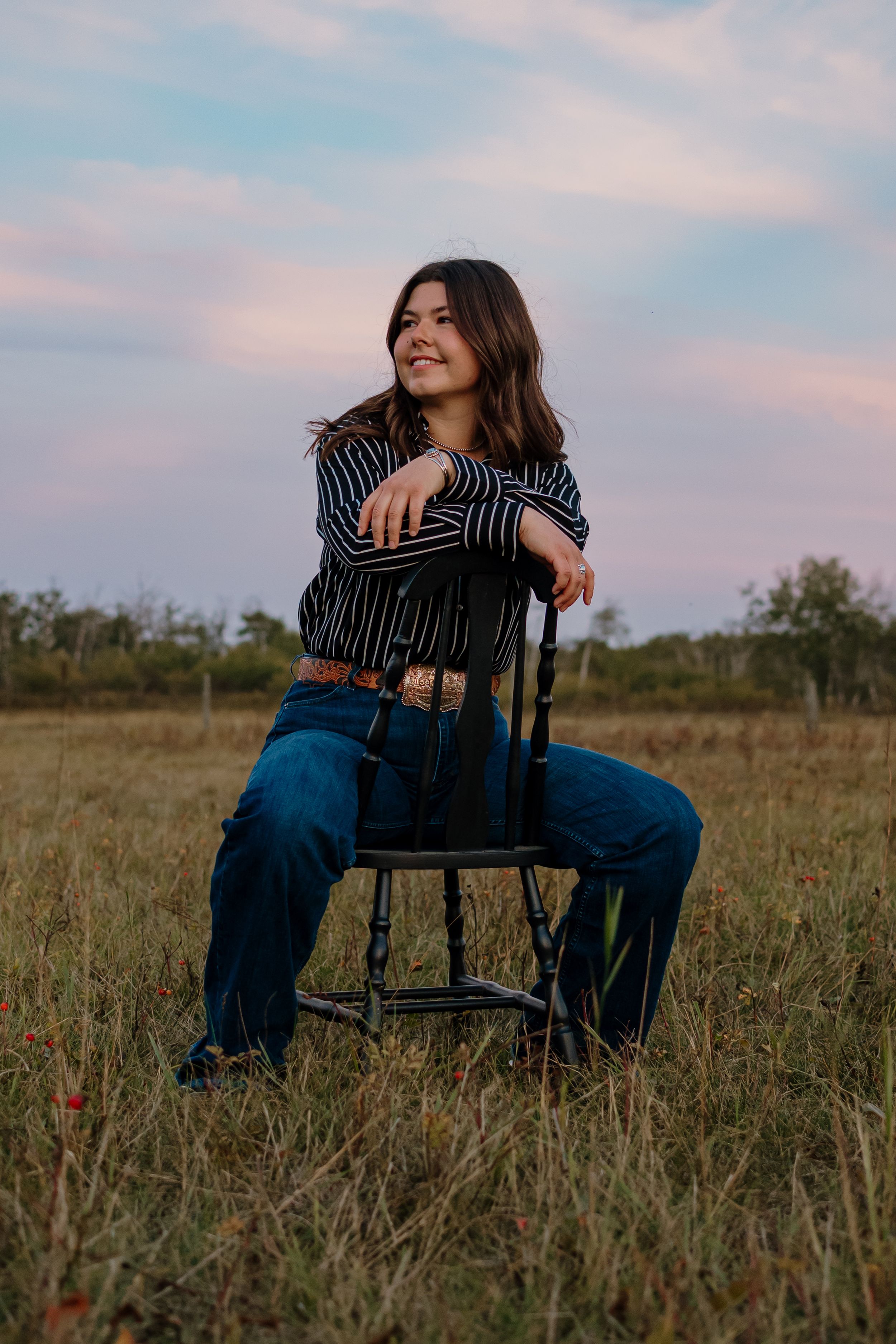 A casual portrait in a striped shirt and jeans sitting on a black chair in a grassy field at sunset.