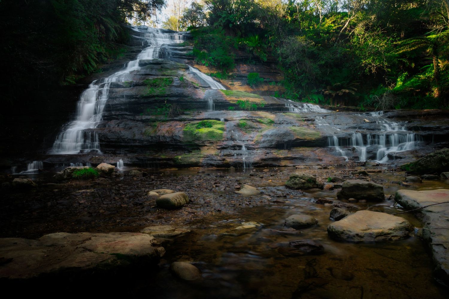 Series of cascading waterfalls flowing over rocky terrain in a lush forest setting.