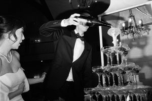 Groom in tuxedo pouring champagne into a tiered glass tower at a black and white wedding reception.