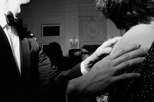 Groom in tuxedo gently placing hand on guests shoulder at dimly lit black and white wedding reception.