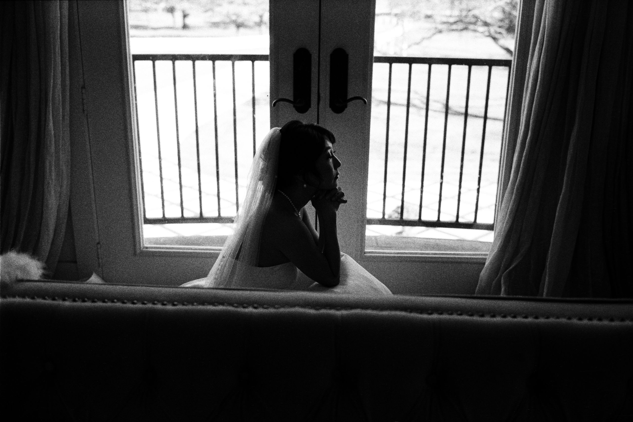 Black and white silhouette of bride in wedding dress sitting by window in contemplative moment.