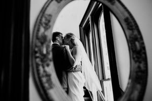 Bride and groom sharing a romantic kiss reflected in an ornate vintage round mirror in black and white.