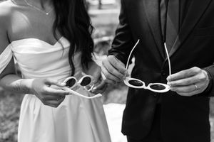 Black and white photo of a bride and groom holding matching white sunglasses outdoors.