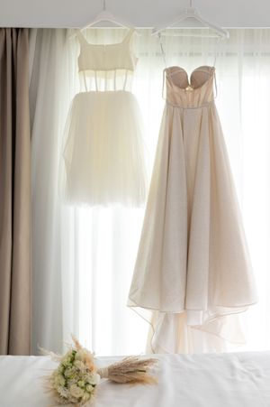 White flower girl dresses and wedding gowns hanging by window with natural light.