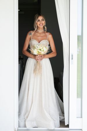 Bride in flowing white wedding gown holding bouquet in doorway with natural lighting.