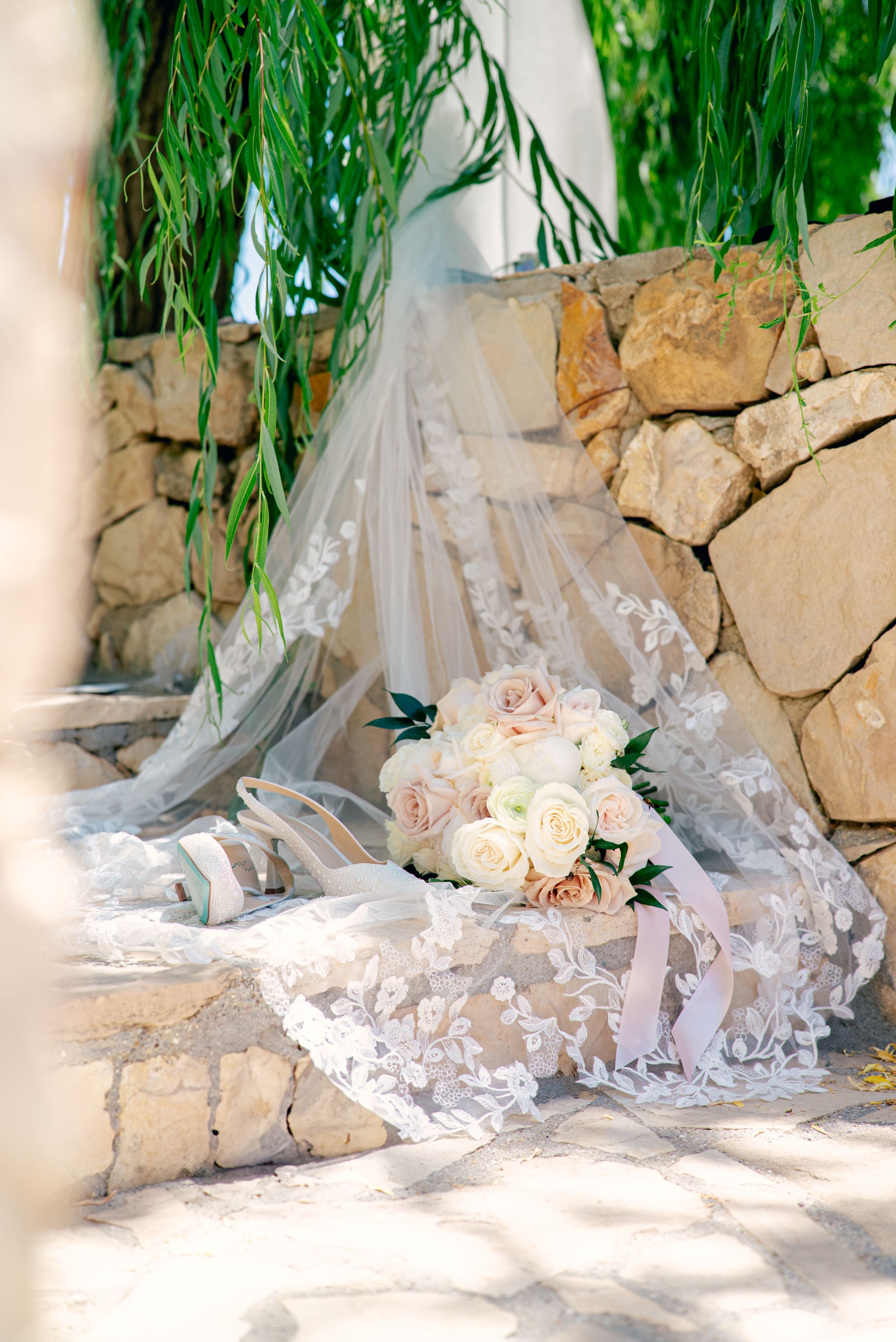 Elegant wedding details including lace veil and blush roses arranged on stone patio under willow branches.
