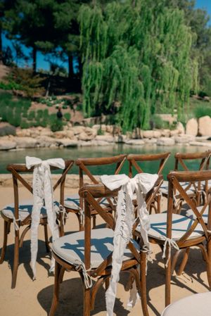 Wooden cross-back chairs with white ribbons set up for an outdoor wedding ceremony by a lake.