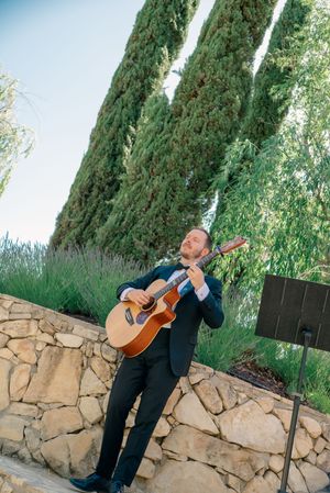 Guitarist performing at an outdoor wedding venue against a stone wall backdrop.