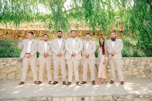 Groomsmen in light beige suits lined up against stone wall under green foliage.