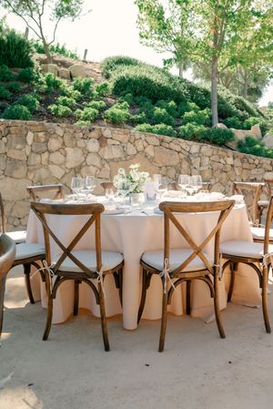 Rustic outdoor wedding reception setup with crossback chairs and white tablecloths.