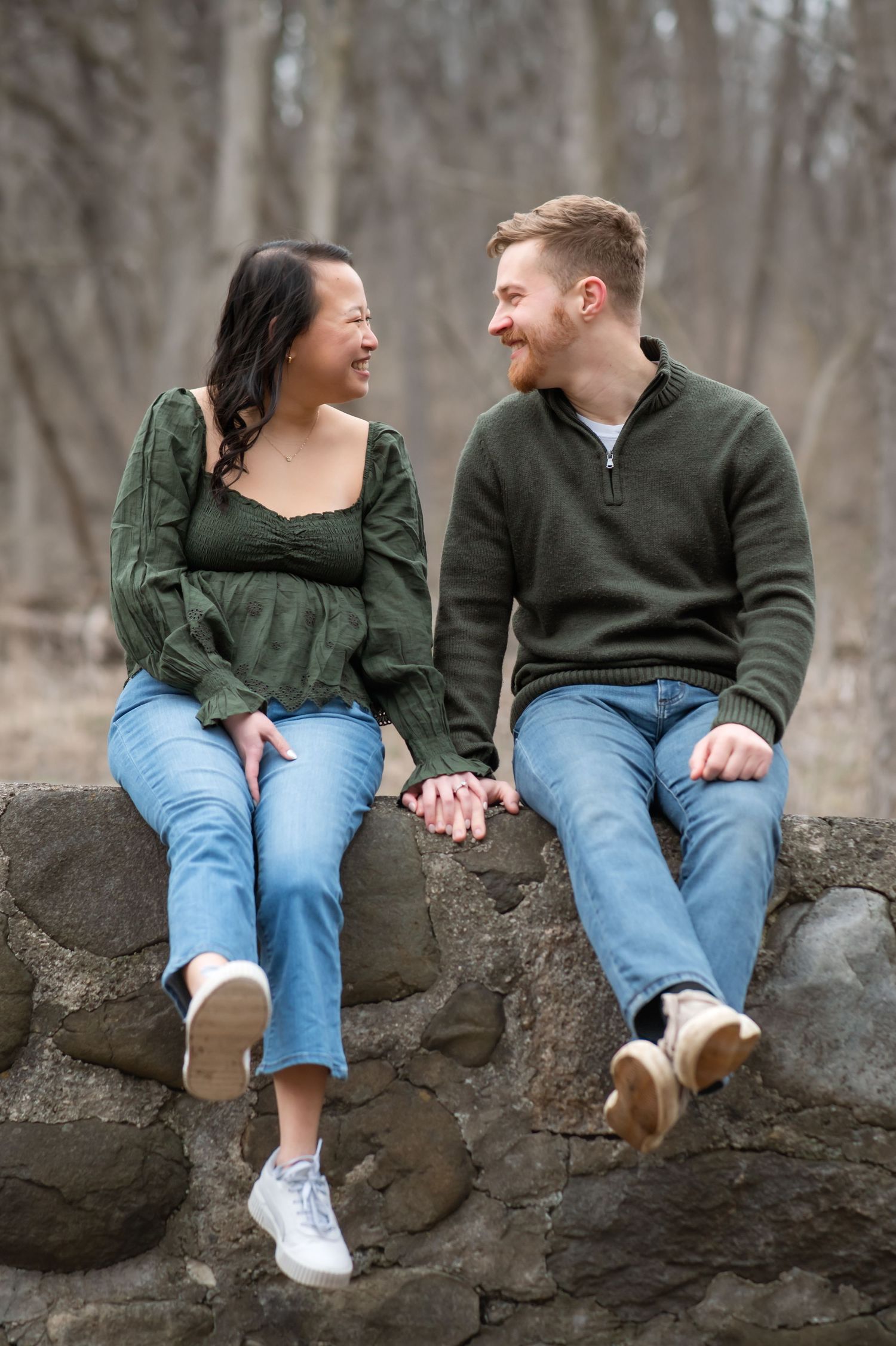 A couple sits on a stone wall wearing matching outfits of jeans and dark sweaters while sharing a candid moment.