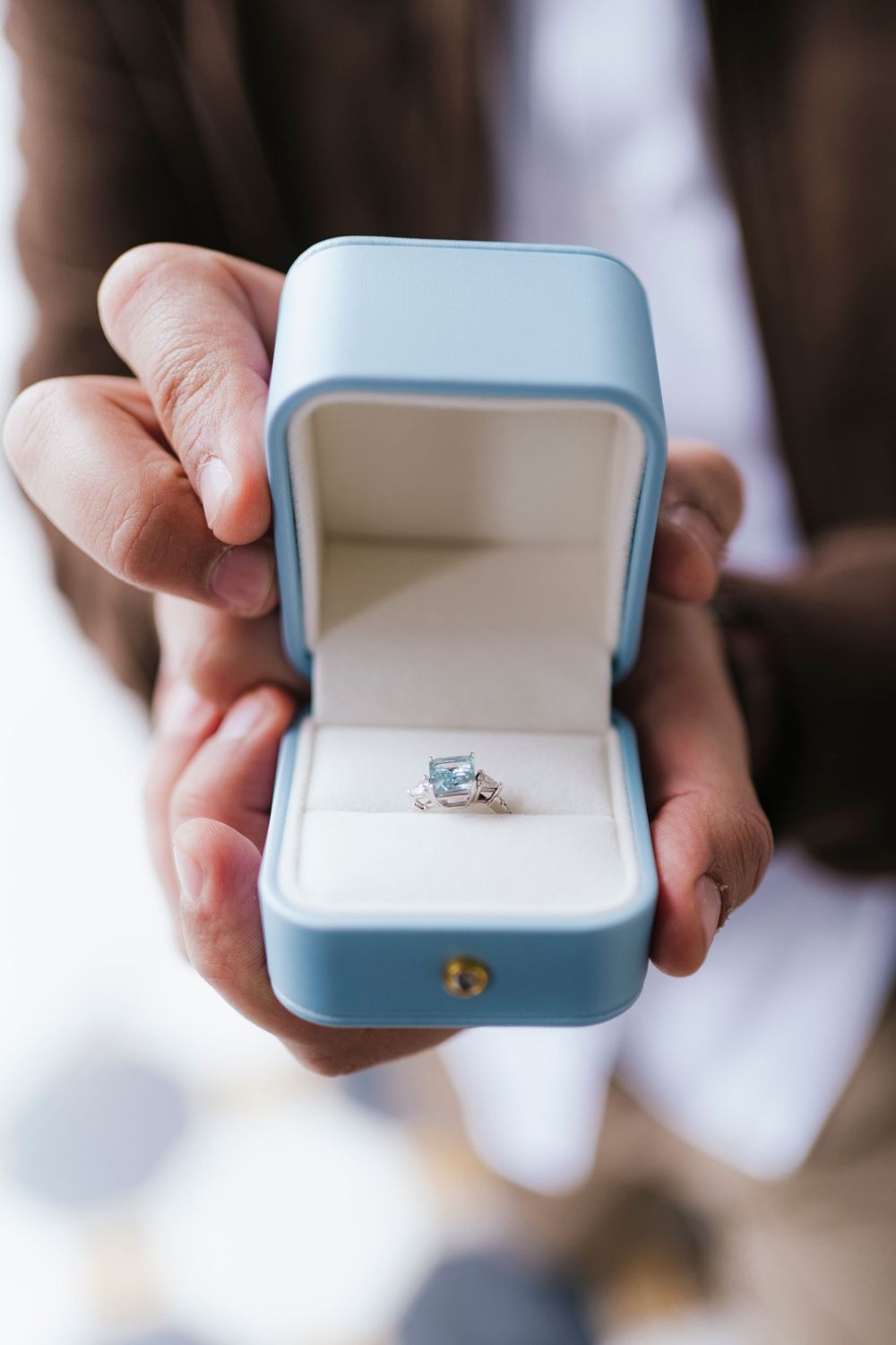 A light blue engagement ring box being held and opened to reveal a sparkling diamond ring.