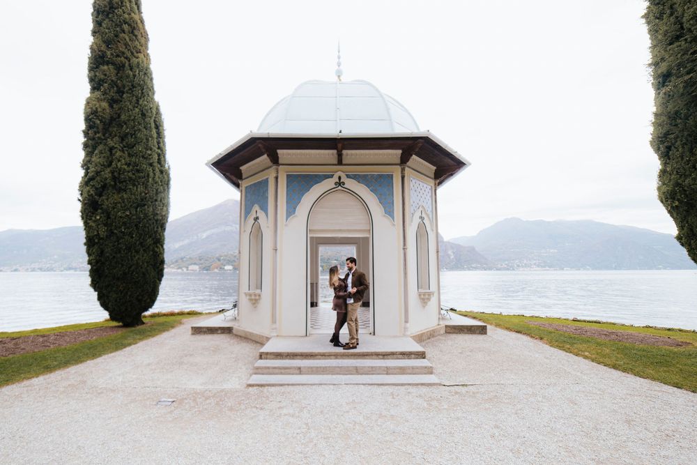 A couple embraces in an elegant white gazebo overlooking a serene lake with cypress trees framing the scene.