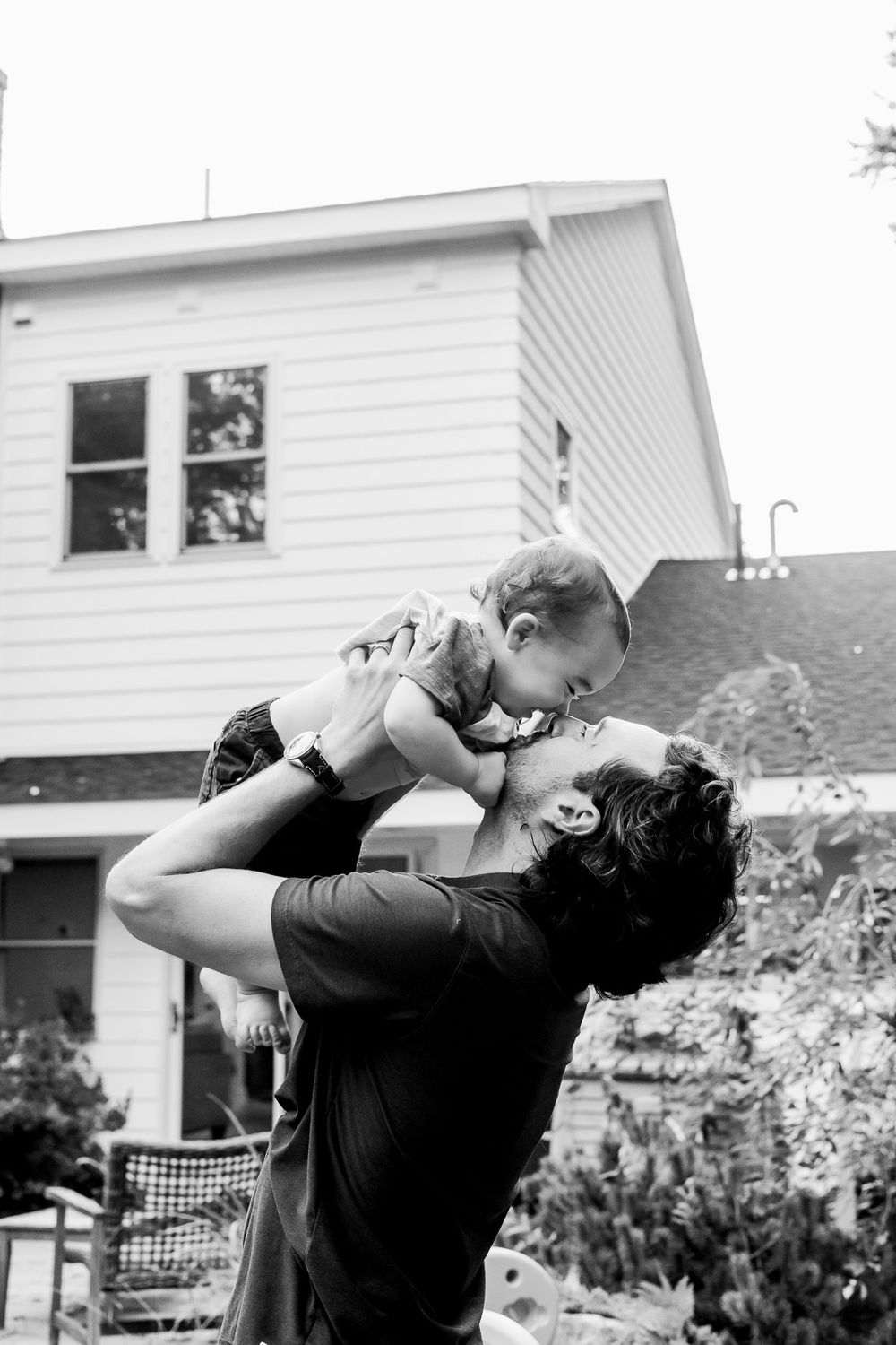 A tender moment captured in black and white showing a parent holding a baby against a white house exterior.