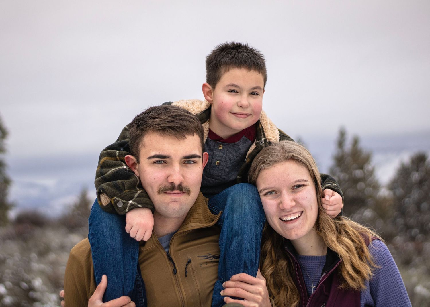 a young man and woman crouch on the ground by younger brother who stands between them and puts his arms on their shoulders. there is snow