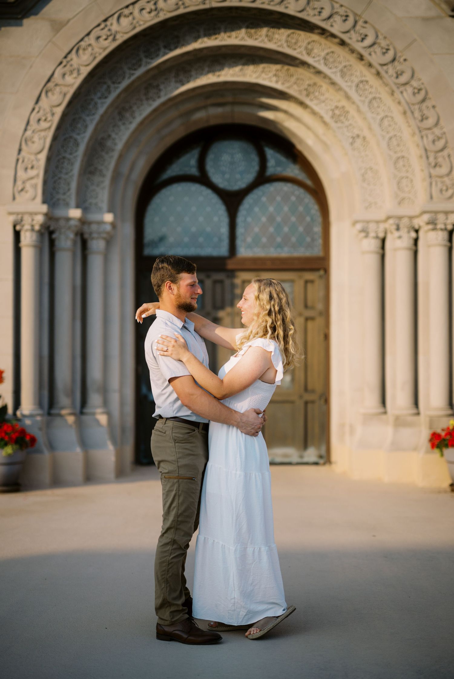 A romantic couple embraces in front of an ornate stone archway with columns and decorative carvings.