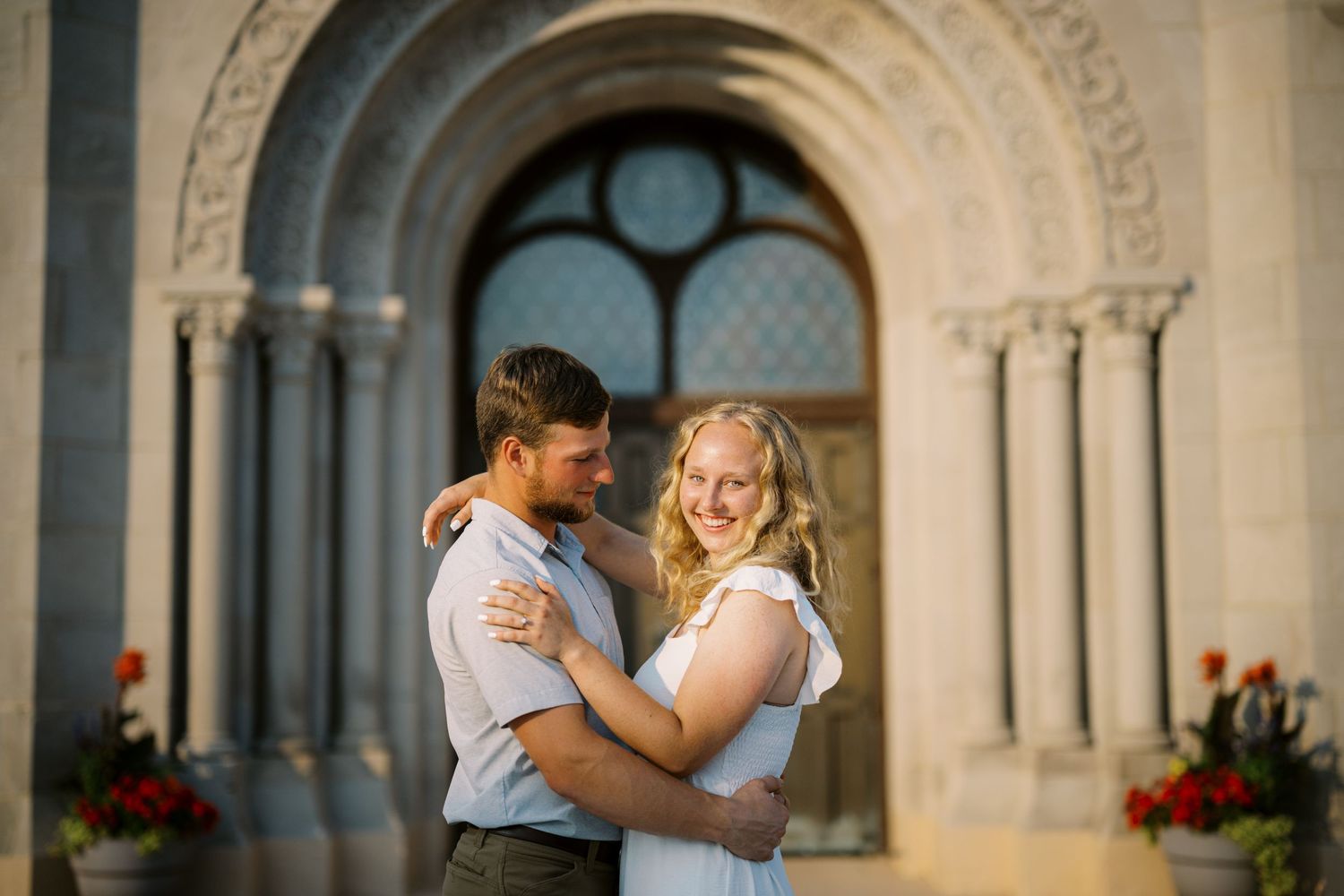 A couple embraces in front of an ornate stone archway with stained glass windows and red flowers.