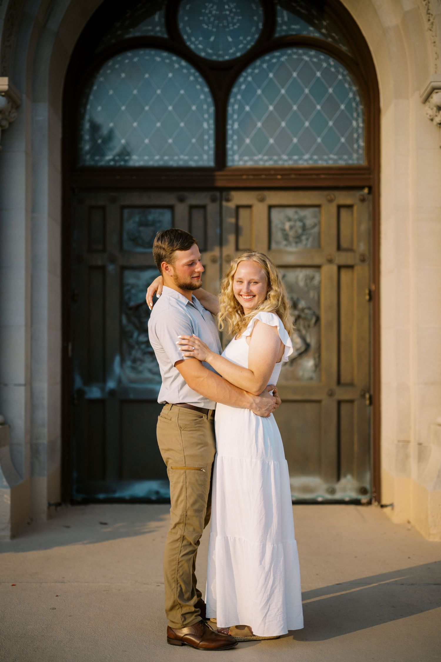 Couple embraces in front of a grand wooden door with arched windows during a golden evening photoshoot.