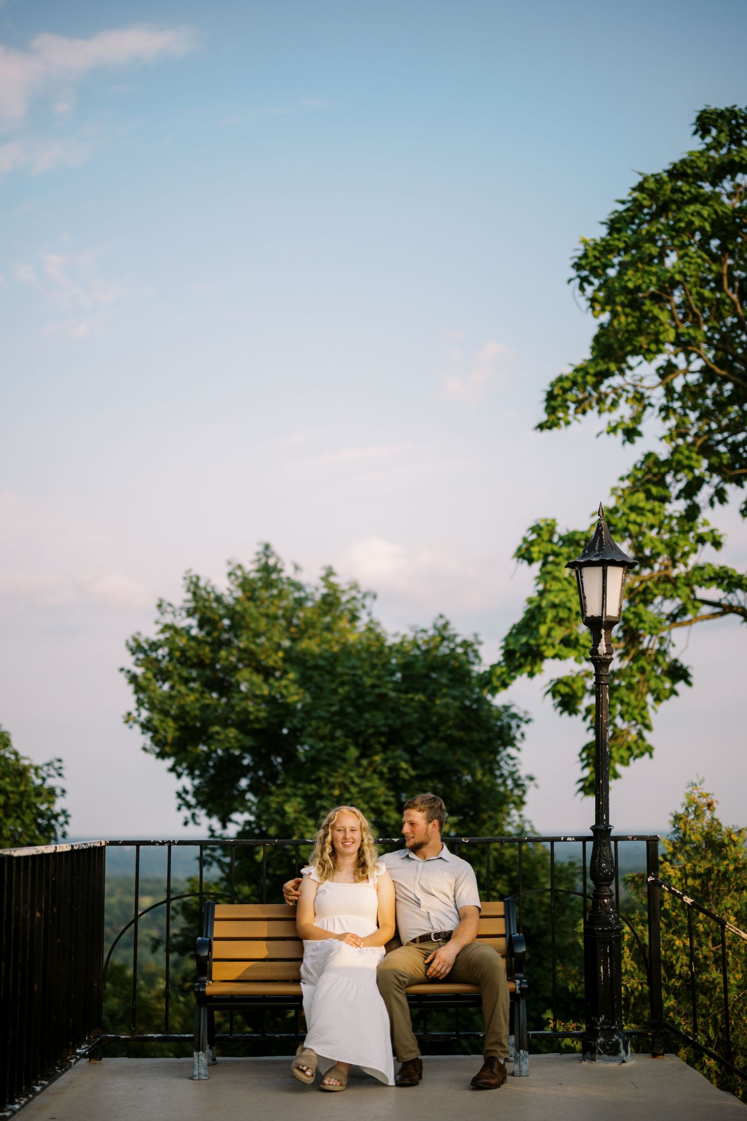 A couple sits together on a wooden bench at sunset overlooking a scenic mountain vista with a lamp post nearby.