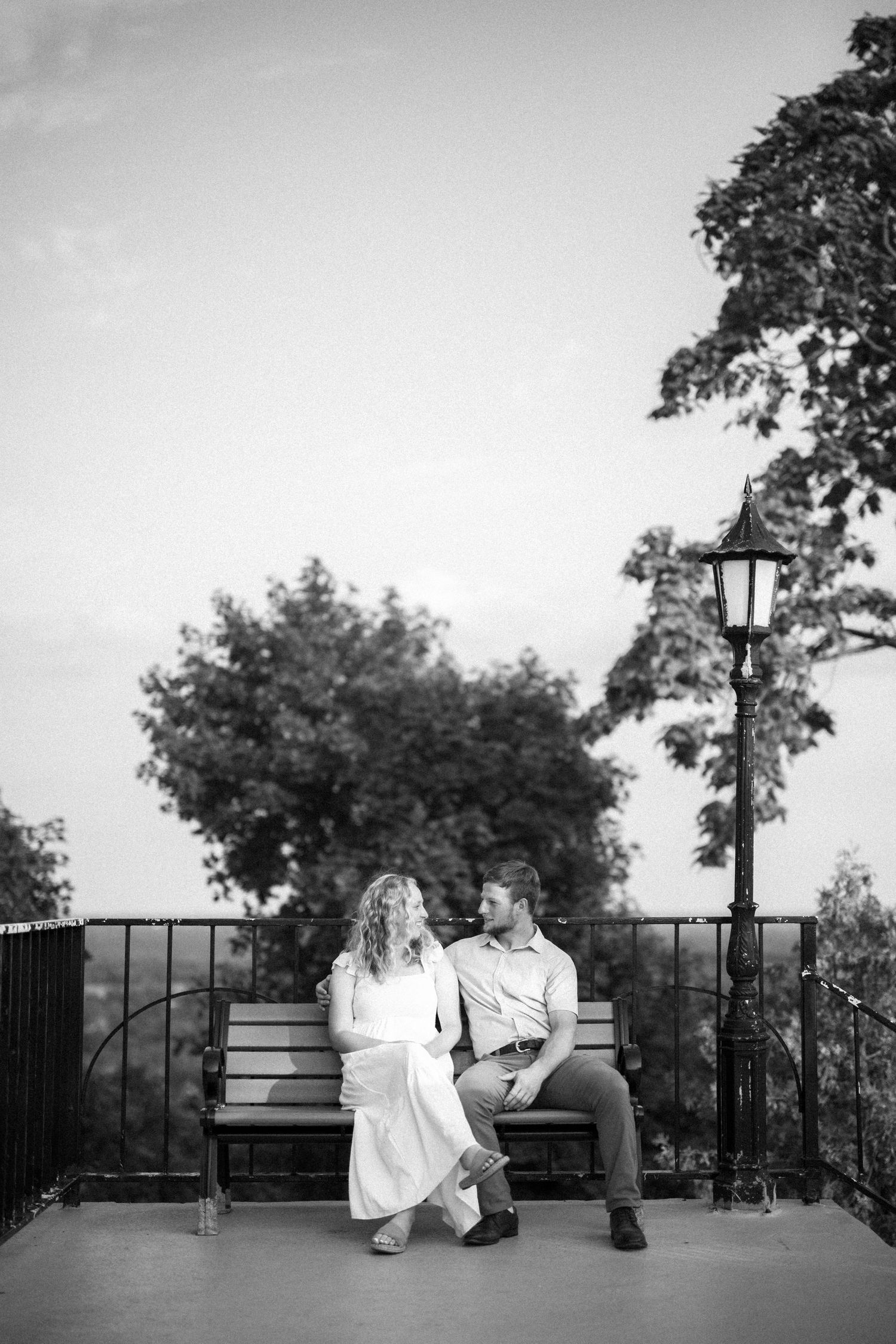 A romantic couple sits together on a bench at dusk with a vintage lantern post and trees in the background.