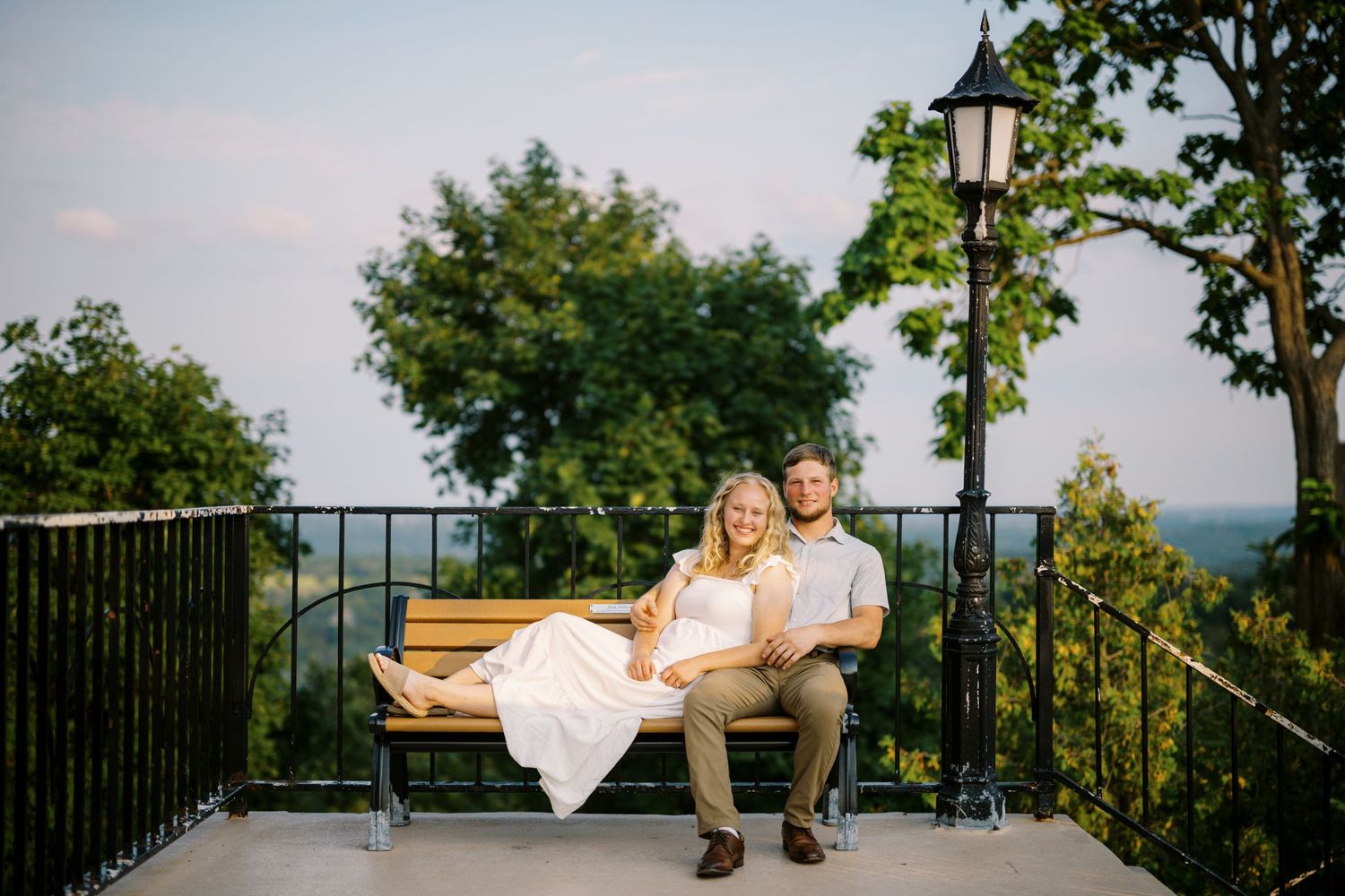 A couple sits on an outdoor bench at sunset overlooking a scenic mountain view with a vintage lamp post nearby.