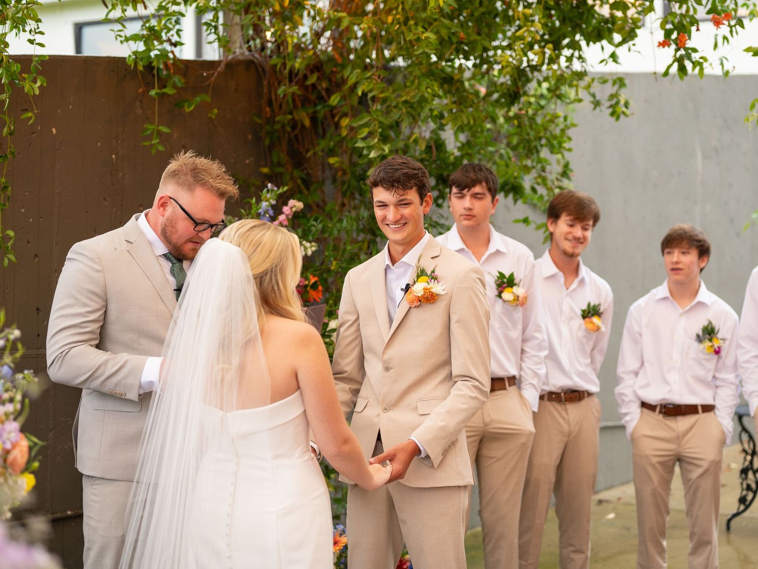 Wedding ceremony moment at outdoor venue with bridal party in khaki suits and bright orange boutonnieres during autumn.