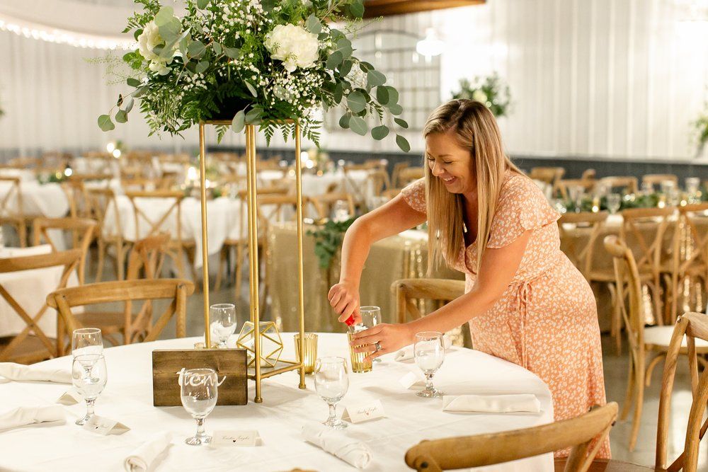 A person in pink arranges elegant table settings with floral centerpieces for a wedding reception.