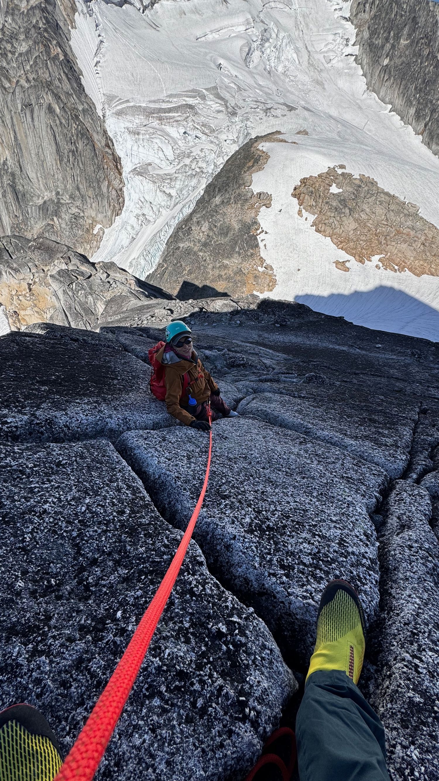 Kristen Lusk ascending dark granite rock face with red rope and snow-covered peaks visible in background in the Bugaboos with Evan Will