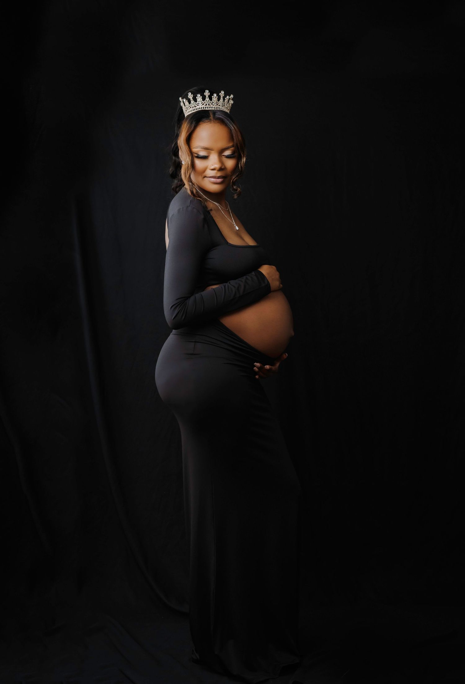 An elegant maternity portrait against a dark background features a glamorous crown and form-fitting black outfit.