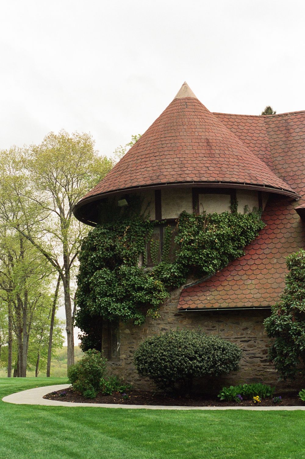 Stone turret building with red tile conical roof, ivy-covered walls, manicured green lawn, and trees in background.