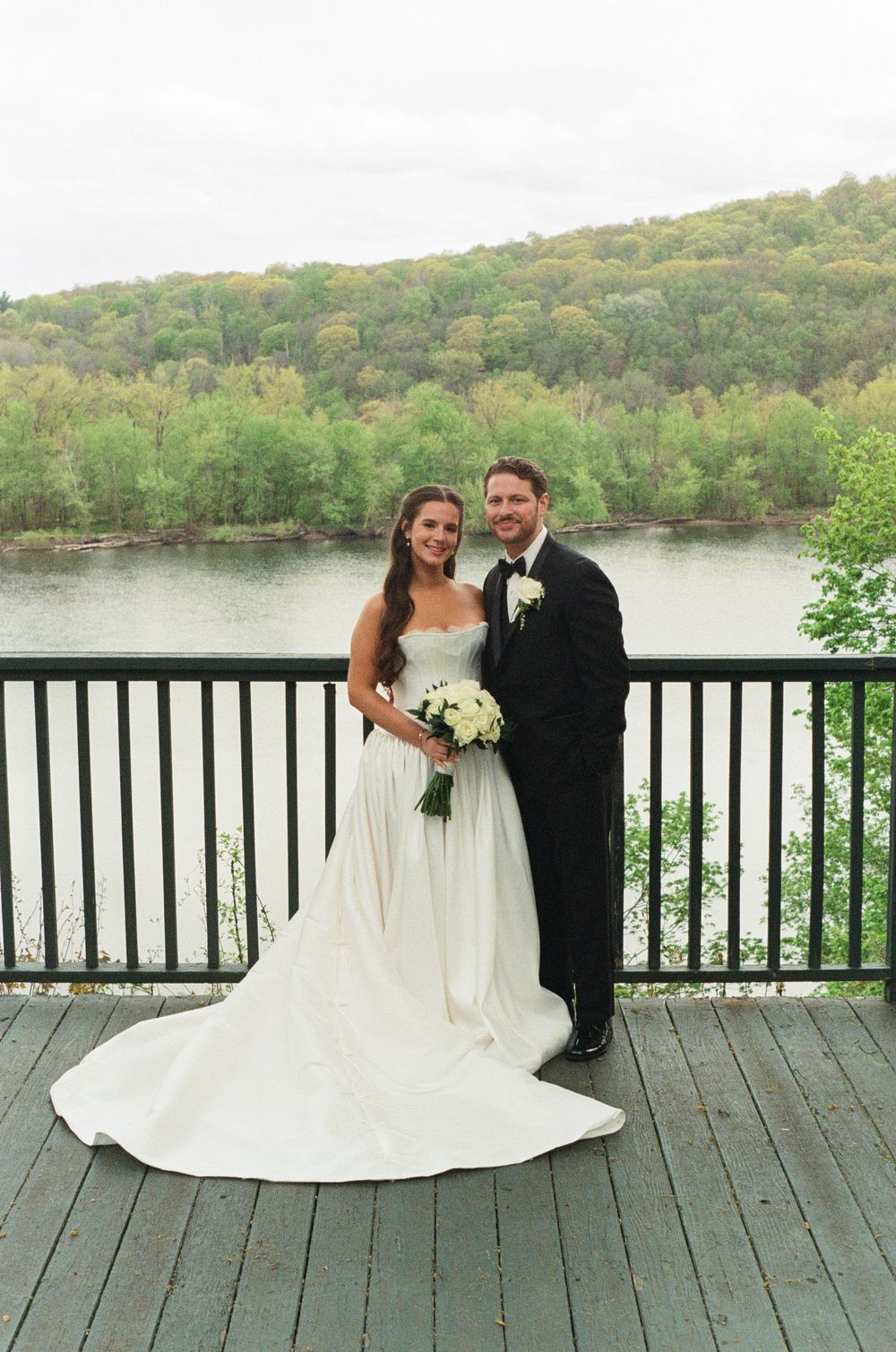 Bride in white gown and groom in tuxedo pose on deck overlooking scenic river and green forested hills.