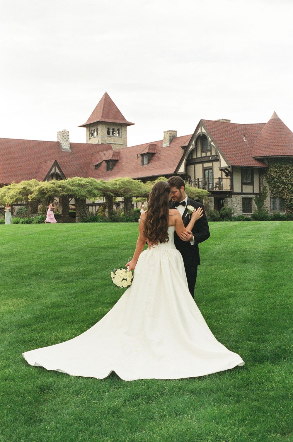 Bride and groom embrace on lush green lawn before elegant Tudor-style estate on their wedding day.