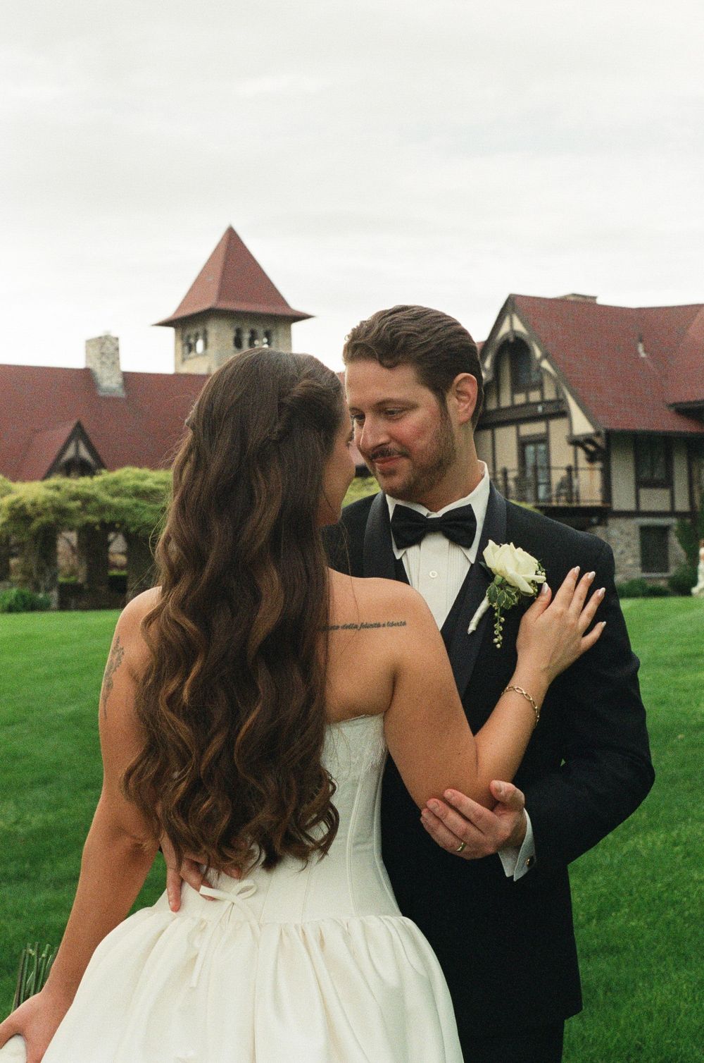 Bride and groom share a romantic moment on green lawn outside Tudor-style venue, bride in strapless gown, groom in black tuxedo.