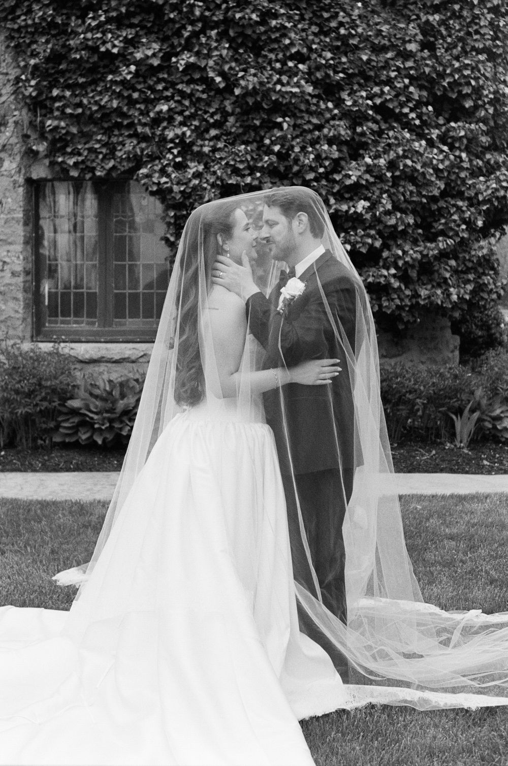 Bride and groom share romantic kiss under flowing veil in black and white wedding portrait outdoors.