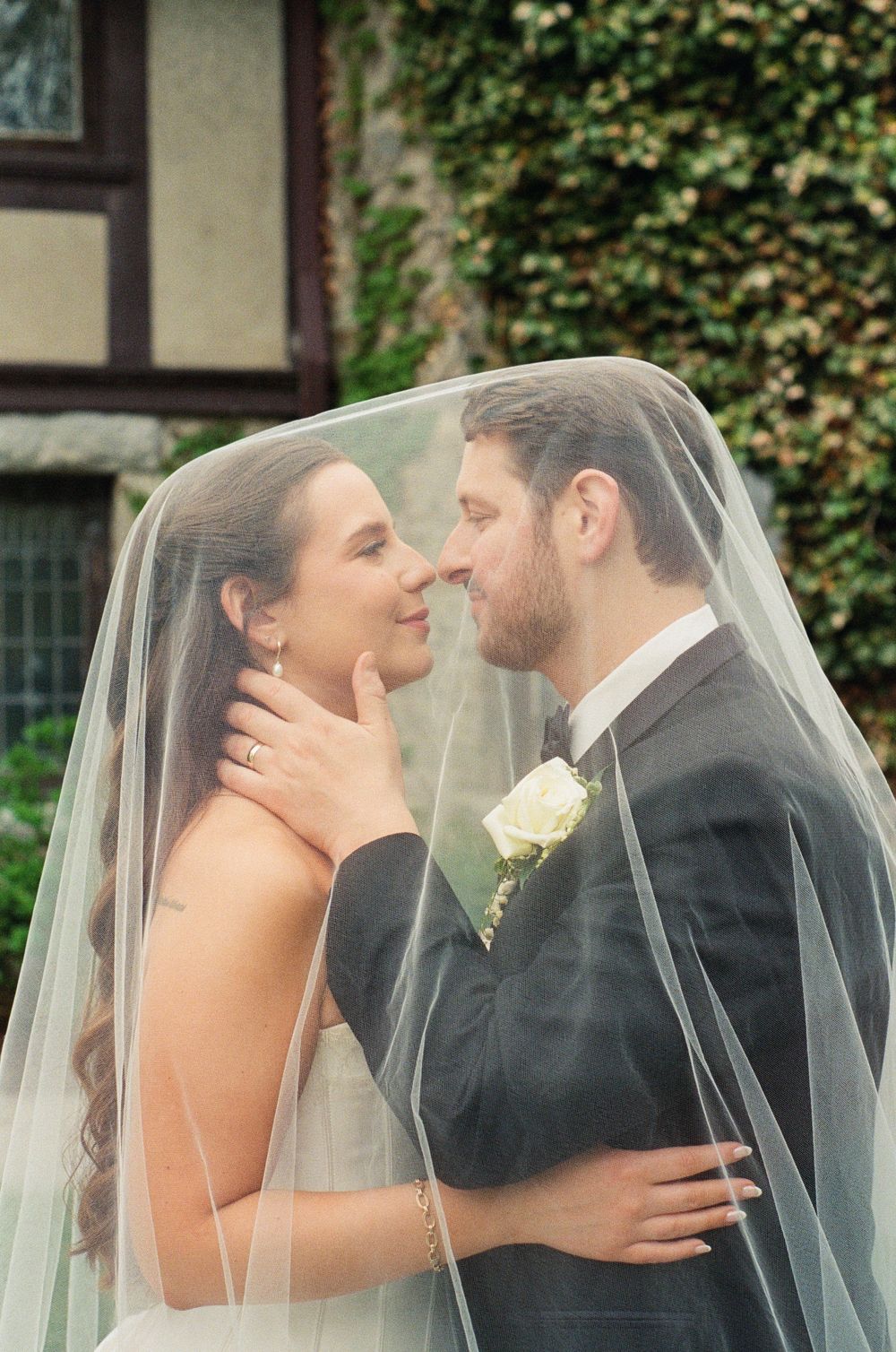Bride and groom share a romantic moment under a wedding veil, nearly kissing outdoors with ivy-covered wall behind them.