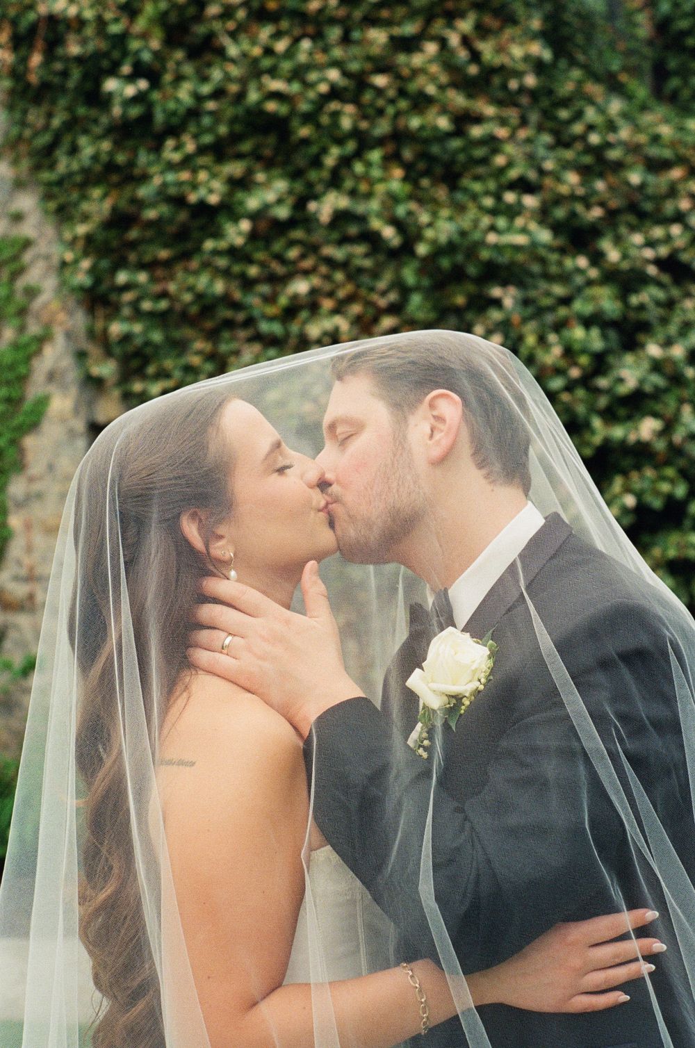 Bride and groom sharing a romantic kiss under a flowing white veil outdoors with lush green ivy wall backdrop.