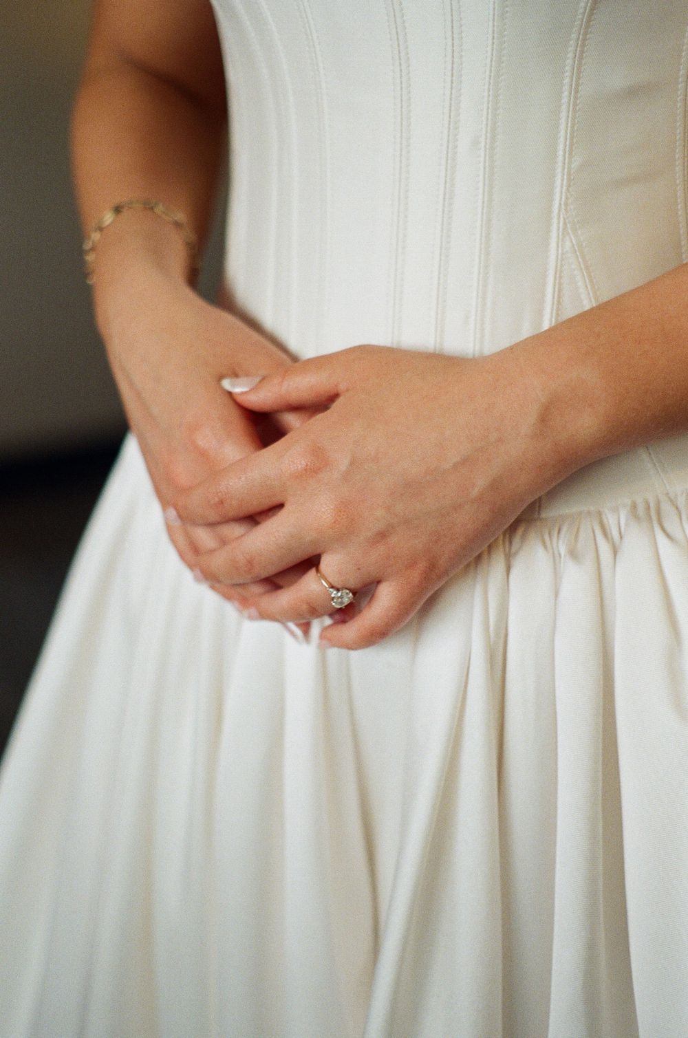 Bride in white wedding gown clasping hands, showcasing diamond engagement ring and gold bracelet.