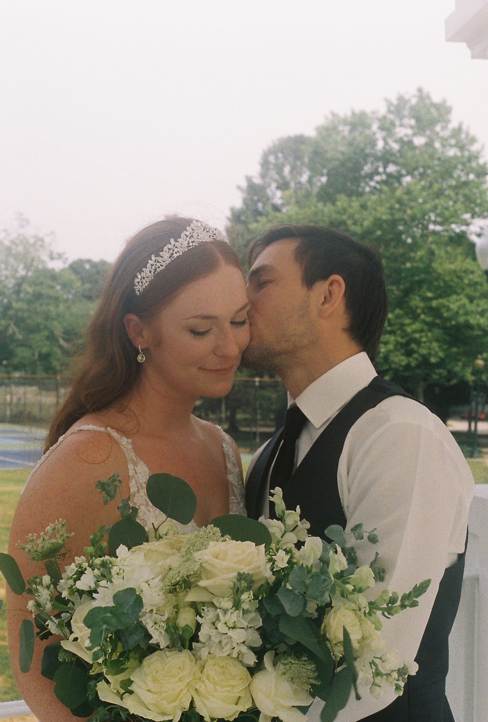 A couple embraces outdoors holding a white and green wedding bouquet.