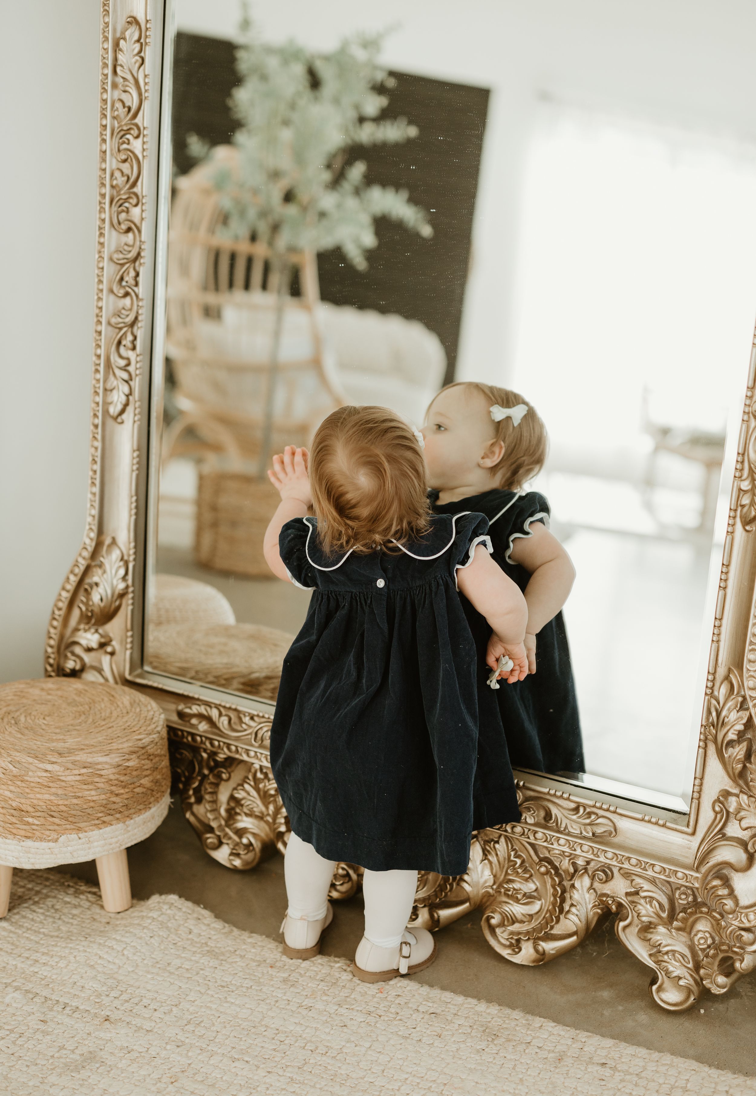 Toddler in black dress looking at reflection in ornate gold mirror against white wall.