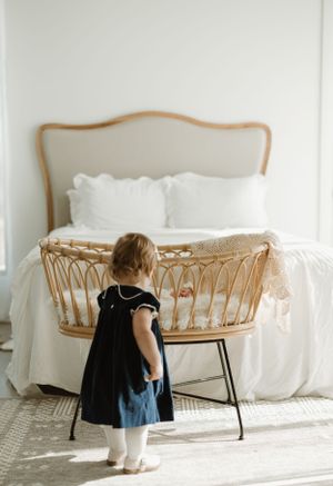 Small child in navy dress looking at woven bassinet in bright white bedroom.