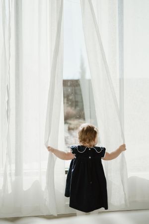 Small child in a dark dress reaching up to touch white curtains by a window.