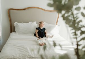 Young child sitting alone on white bedding against an upholstered headboard.