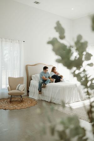 A cozy bedroom scene with a white bed, beige armchair, and sunlit window with green plant shadows.