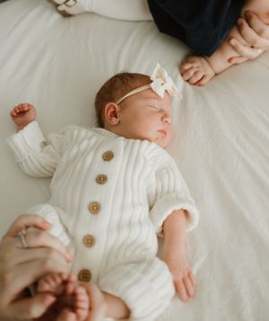 Sleeping infant wearing white knit outfit with wooden buttons on white bedding.