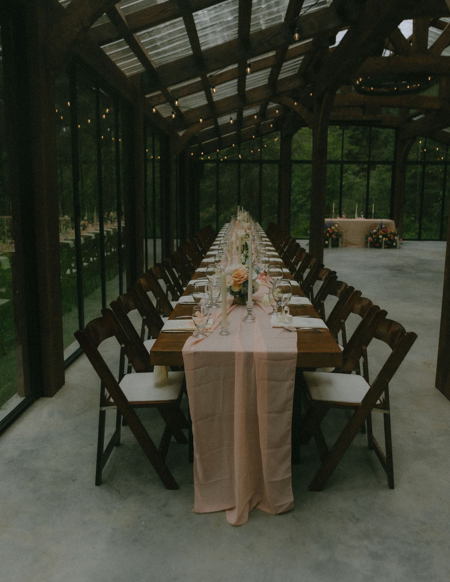 An elegant wedding reception table setting in a rustic greenhouse with wooden chairs.
