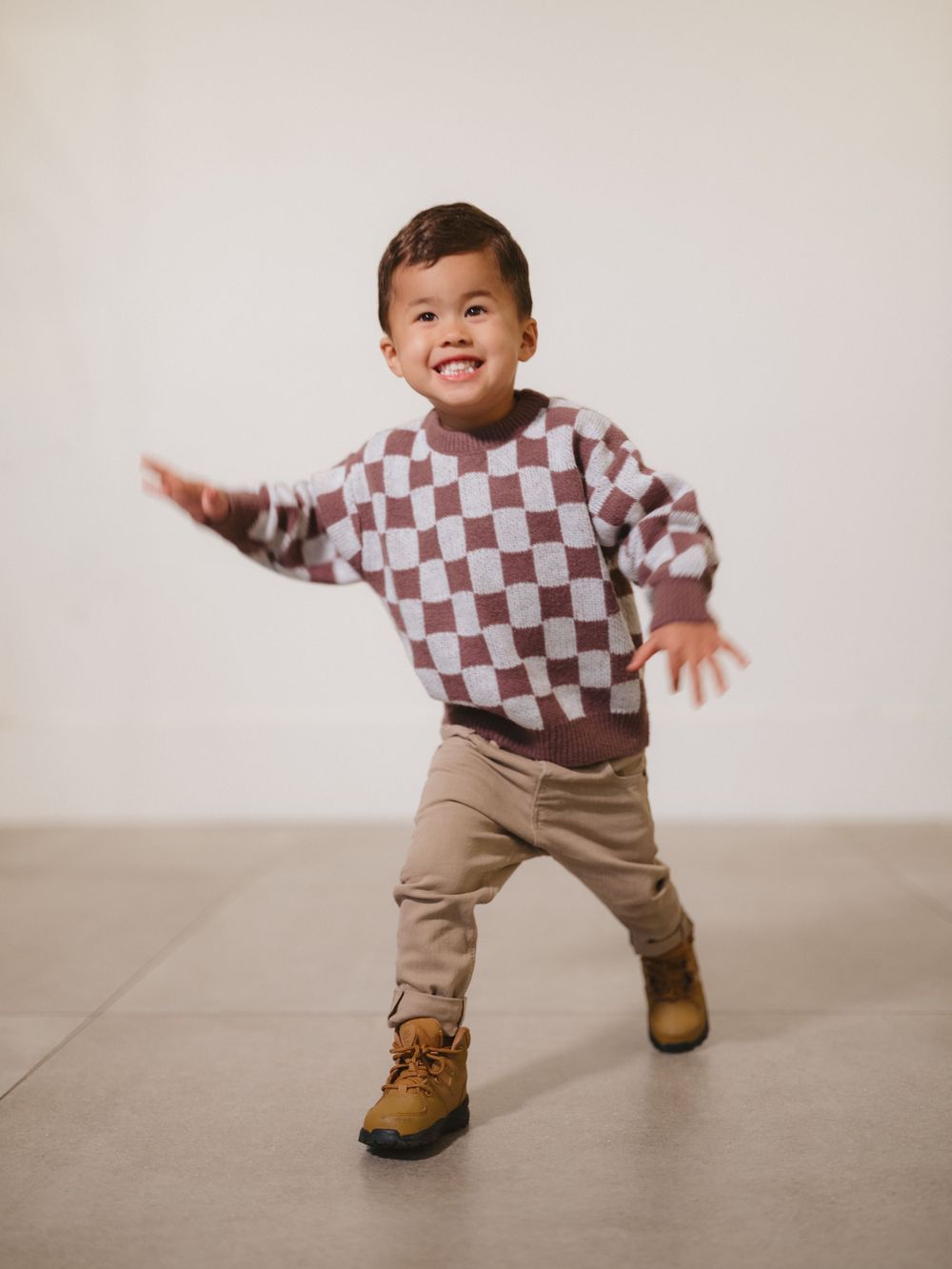A child wearing a checkered sweater and khaki pants strikes a playful dance pose against a neutral background.