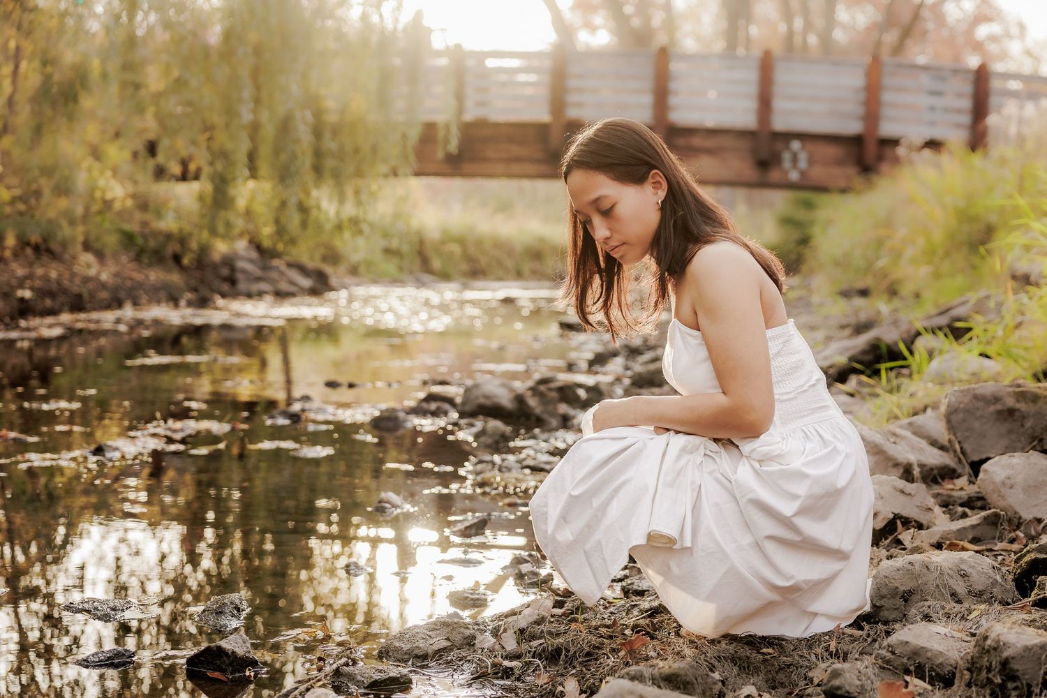 A person in a white dress crouches by a stream among rocks on a sunny day.